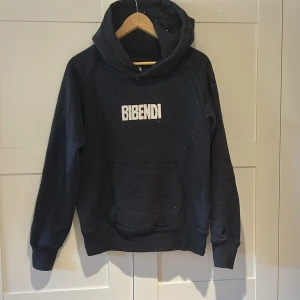 Svart hoodie Bibendi RedLine S - Svart hoodie från RedLine i storlek S med broderad Bibendi-text på bröstet och en cool vit tryckt figur på ryggen som får hoodien att sticka ut på ett mycket snyggt sätt. Klassisk känguruficka framtill och stor huva. Perfekt för dig som gillar streetstyle och vill sticka ut lite extra.