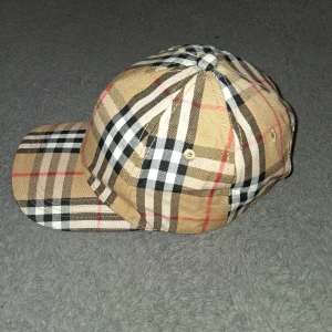 Burberry rutig keps i bomull - Snygg keps från Burberry i klassiskt beige rutigt mönster med svarta, vita och röda detaljer. Justerbar rem med guldfärgad spänne bak och broderad Burberry-logga på insidan. Tillverkad i bomull, perfekt för en trendig streetstyle.