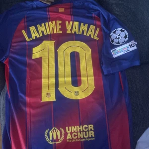 Barcelona Lamine Yamal tröja  - Snygg FC Barcelona matchtröja med Lamine Yamal och nummer 10 på ryggen. Tröjan är blå och röd med gula detaljer, randigt mönster och officiella klubbmärken. Materialet är lätt och andas, perfekt för fotboll. UEFA Foundation-märke på ärmen och Spotify-logga på bröstet. storlek M ny och oanvänd 