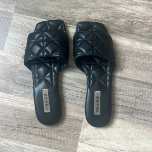 Svarta quiltade sandaler från NA-KD - Stilrena svarta sandaler från NA-KD med bred quiltad rem och fyrkantig tå. Tillverkade i syntetmaterial med mjuk vaddering för extra komfort. Perfekta för en trendig look under varma dagar.