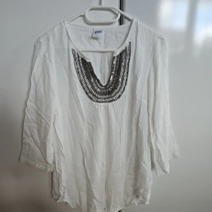 Vit blus med paljettdekor från vero moda - Vit blus från YERB med snygg paljettdekor i silver runt den djupa v-ringningen. Blusen har trekvartsärm och är gjord i ett lätt och luftigt bomullstyg. Perfekt för dig som gillar att sticka ut med detaljer och vill ha en chill men ändå cool look.