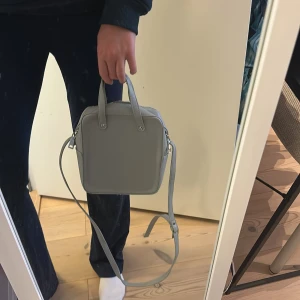 Ljusgrå fyrkantig axelväska - Säljer en stilren ljusgrå axelväska med fyrkantig form. Väskan har både handtag och justerbar axelrem samt dragkedja upptill. I väskan finns ett fack med dragkedja för småsaker och även ett fack utan dragkedja. Den är använd endast ett par fåtal gånger, väldigt bra skick. Säljer pga att den inte kommer till användning😊💕