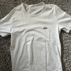 Vit t-shirt från BOSS - Klassisk vit t-shirt från BOSS med diskret svart logga broderad på bröstet. T-shirten har rund halsringning och korta ärmar. Tillverkad i mjuk bomull för en skön och avslappnad känsla.
