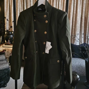 Mörkgrön skepparkavaj från Zara - Stilren mörkgrön skepparkavaj från Zara med dubbelknäppning och guldiga knappar framtill och vid ärmslut. Jackan har hög krage, två fickor med lock och markerad midja för en snygg siluett. Perfekt för dig som vill ha en klassisk men edgy look.