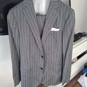 Grå randig kavaj med vita ränder - Snygg grå kavaj med vita vertikala ränder och klassisk passform. Kavajen har två knappar framtill, fickor med lock och en bröstficka med vit näsduk. Perfekt för en stilren och modern look komplett med väst och byxa