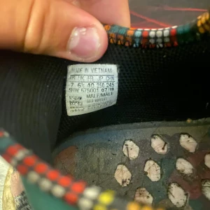 Adidas Pharrell Williams Hu NMD Moth3r L4nd - Unika Adidas Pharrell Williams Hu NMD sneakers med texten 'MOTH3R L4ND' på ovandelen. Skorna har färgglada detaljer i blått, rosa, gult och grönt samt stickat mönster och vit sula. Perfekt för dig som vill sticka ut med stil och komfort.