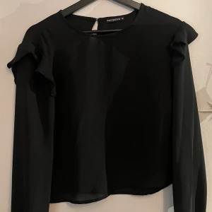 Svart blus med volang från Terranova - Svart blus från Terranova med långa ärmar och volangdetaljer på axlarna. Tunn och luftig känsla, Rund halsringning och enkel knäppning bak i nacken.