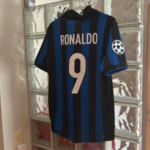 Inter Ronaldo 9 fotbollströja Nike M - Säljer en klassisk Inter Milan fotbollströja med Ronaldo 9 på ryggen. Tröjan är blå och svart med vertikala ränder, Champions League-märke på ärmen och Pirelli som sponsor framtill. Ikonisk gul Nike-logga och klubbmärke med stjärna. Perfekt för samlare eller fans!