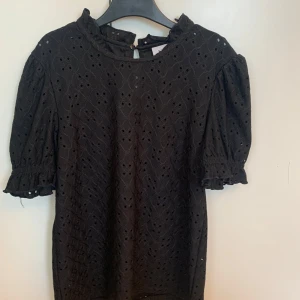 Svart blus med broderade detaljer från Vila - Supersöt svart blus från Vila i storlek XS med broderade hålmönster och volangdetaljer vid ärmslut och hals. Blusen har korta puffärmar och knäppning bak i nacken. 