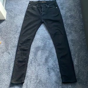Svarta jeans från Nudie Jeans - Svarta jeans från Nudie Jeans i modellen 29/32. Jeansen har klassisk femficksdesign, svarta sömmar och Nudie-logga på bakfickan. Snygg passform med smala ben och midja med hällor för bälte. Perfekta för en clean och stilren look.