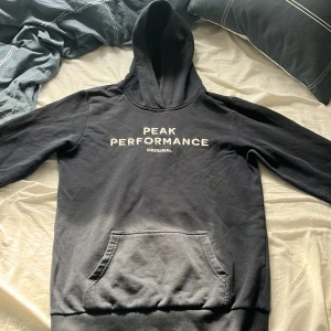 Mörkblå hoodie Peak Performance 170 - Svart hoodie från Peak Performance med vit broderad logga på bröstet. Klassisk känguruficka framtill, huva och långa ärmar. Mjuk och skön, perfekt för chill eller skola. Snygg och enkel design som funkar till det mesta.