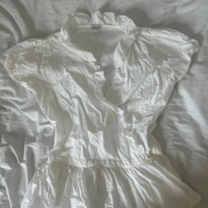 Vit omlottblus från Hunkydory - Supervacker, exklusiv vit omlottblus från Hunkydory Essentials i 100% bomull! Blusen har omlott-skärning, v-ringning och korta ärmar med volangdetaljer. Perfekt för en klassisk look och ger en fräsch och luftig känsla.