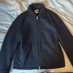 Svart ulljacka från COS, storlek M - Säljer en svart jacka från COS i storlek M. Jackan är i ullmaterial, har rak passform och stängs med dragkedja. Klassisk krage och två snedställda fickor framtill. Perfekt för dig som gillar stilren och enkel design.