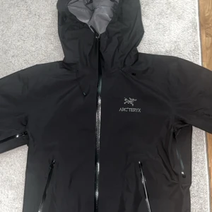 Svart Arc'teryx vindjacka med huva - Säljer en svart vindjacka från Arc'teryx i storlek L. Jackan har huva, vattentäta dragkedjor och minimalistisk design. Perfekt för blåsiga dagar och outdoor-äventyr. Materialet är lätt och känns riktigt smidigt. Snygg logga på bröstet och flera praktiska fickor.