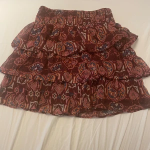 Mönstrad volangkjol från Lindex - Supersöt kort volangkjol från Lindex i vinröda och rosa toner med paisleyinspirerat mönster. Kjolen har resår i midjan och flera lager volanger som ger en flowig känsla. Perfekt för dig som gillar färg och mönster!