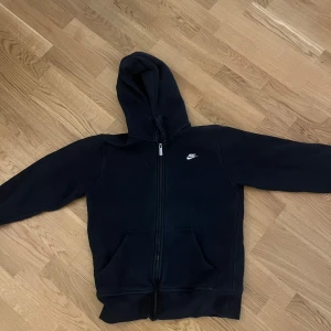 Svart Nike hoodie med dragkedja - Svart hoodie från Nike med broderad logga på bröstet. Tröjan har huva med snörning, hel dragkedja och två fickor framtill.