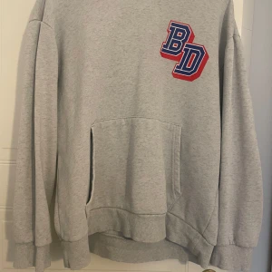 Grå hoodie med BD-tryck - Snygg grå hoodie med stora BD-tryck i blått och rött både fram och bak. Perfekt för dig som gillar sportig och avslappnad stil.  Långärmad och har en loose, oversize fit.
