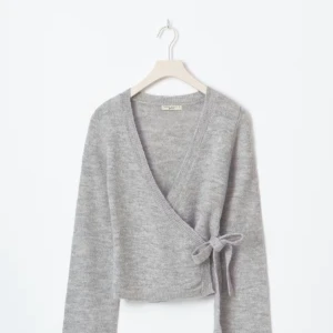 Grå omlottkofta med knyt från Gina Tricot  - Supermjuk grå omlottkofta från Gina Tricot med knyt i sidan och v-ringad hals. Koftan är stickad, har långa ärmar och en croppad passform som ger en chill vibe. Perfekt att slänga över ett linne eller t-shirt för en snygg lager-på-lager-look.