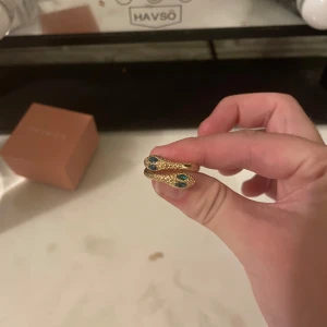 Guldig ormring med gröna stenar - Cool ring i form av en orm, i guldig färg med detaljerade fjäll och gröna stenar som ögon. Ringen har en unik och trendig design som verkligen sticker ut och passar perfekt för dig som gillar statement-smycken.