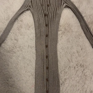 Glittrig ribbad långklänning Rita Ora - Supersnygg långklänning från Rita Ora x Primark i glittrigt ribbat material. Klänningen är brun med guldiga inslag, har långa ärmar, v-ringning och knappar hela vägen framtill. Perfekt för dig som vill sticka ut med stilren glamour. Aldrig använt 