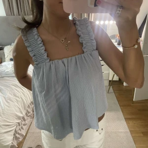 Ljusblå ärmlös linnetopp med volangband - Supersöt ljusblå linnetopp med breda volangiga axelband och luftig passform. Toppen har en fyrkantig ringning och kan stylas med eller utan knytband i midjan för olika looks. Perfekt för varma dagar och ger en fräsch vibe till outfiten.