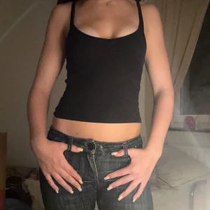 Svart ribbad linnetopp - Säljer en svart ribbad linnetopp med smala axelband och croppad passform. Toppen har en djup urringning och sitter tight mot kroppen, perfekt att styla med jeans eller kjol.