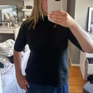 Svart stickad polotröja med kort ärm - Svart stickad polotröja med korta ärmar och lös passform. Tröjan har en hög, vikt krage och är perfekt för lager-på-lager. Enkel och stilren design som funkar till många olika outfits.