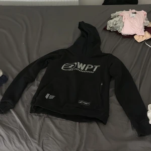Svart WPT hoodie World Padel Tour - Svart hoodie från World Padel Tour med stor WPT-logga på bröstet och tryck vid fickan. Klassisk huva, långa ärmar och ribbade muddar. Perfekt för dig som gillar sportig stil och padel. Mjuk och skön, troligtvis i bomullsmix.