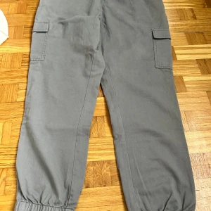 Grå cargobyxor med resår - Snygga grå cargobyxor med elastisk midja och muddar vid benslut. Byxorna har stora sidofickor med lock och är tillverkade i ett mjukt bomullstyg. Perfekta för en avslappnad och trendig streetstil.