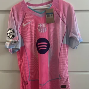 Barça x Nike rosa matchtröja De Jong - Säljer en FC Barcelona matchtröja från Nike i rosa och ljusblått med vita detaljer. Tröjan har korta ärmar, Dri-FIT ADV-material och tryck med F. De Jong 21 på ryggen. Champions League-märke på ärmen och Spotify-logga på bröstet. Perfekt för dig som älskar fotboll och stil.