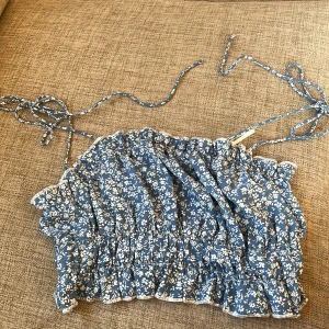Blå blommig croppad linnetopp - Supersöt croppad linnetopp i blått med vitt blommönster och smockad resår. Toppen har tunna axelband med knyt och volangdetaljer längs kanterna. 