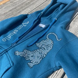 Blå hoodie med strasstiger från Vonstudio - Blå hoodie från Vonstudio med dragkedja, huva och snörning. På bröstet finns en stor tiger i glittrande strass och texten 'Vonstudio' i strass längs dragkedjan. Mjuk insida och känguruficka framtill. Perfekt statement-plagg för dig som gillar unika detaljer.
