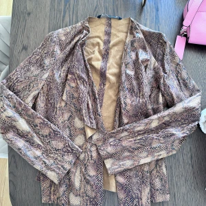 Ormmönstrad mockajacka i brunt - Säljer en snygg jacka med ormmönster i olika bruna och beige toner. Jackan har öppen front och är tillverkad i mockaliknande material. Perfekt för dig som vill sticka ut med en trendig look. Långärmad och loose fit.