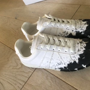Maison margiela paint splatter  - Maison Margiela sneakers i vitt tyg och skinn med unik svart färgstänksdetalj. Klassisk siluett med snörning och platt sula. Kommer med originalkartong. Perfekta för dig som gillar statement och streetstyle. As feta för sommaren och passar till mycket, priset är inte fast, så kom med bud✅