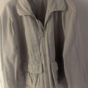 Beige vindjacka med dragkedja från hope  - Snygg beige vindjacka med klassisk krage och dragkedja framtill. Jackan har två stora fickor med lock och en enkel, stilren design. Perfekt för dig som gillar en clean och avslappnad look. Passar bra till jeans eller cargo pants.