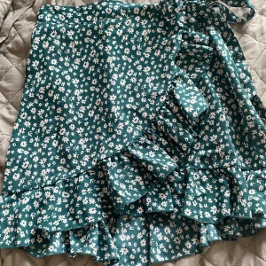 Grön blommig omlottkjol från SHEIN - Supersöt grön kortkjol från SHEIN med vitt blommönster. Kjolen har omlottmodell med volangkant och knyt i midjan, vilket ger en riktigt gullig och somrig vibe. Perfekt för dig som gillar en feminin och trendig stil!