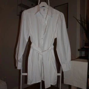 Vit skjortklänning med bälte - Vit kort skjortklänning med klassisk krage, långa ärmar och knappar framtill. Klänningen har ett matchande bälte i midjan för snygg passform. Perfekt för en clean och stilren look.