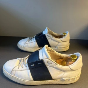 Valentino Garavani Open sneakers vit/blå - Snygga Valentino Garavani Open sneakers i vitt skinn med bred marinblå läderdetalj över mitten. Klassisk rund tå, snörning och ikoniska nitar baktill på sulan. Perfekta för dig som gillar exklusiva och stilrena sneakers med en twist.