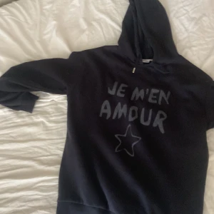 Svart hoodie med text och stjärna - Svart hoodie med huva och trycket 'JE M'EN AMOUR' i grått framtill samt en handritad stjärna under texten. Tröjan har långa ärmar och en klassisk, avslappnad passform. Perfekt för dig som gillar statement-plagg med fransk vibe.