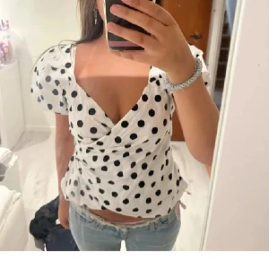 Prickig omlottopp  - Superfin vit omlottopp med svarta prickar. Toppen har puffiga korta ärmar, v-ringning och markerad midja. Perfekt för dig som gillar en söt och trendig look. Materialet är mjukt och lätt, passar perfekt till jeans eller kjol.