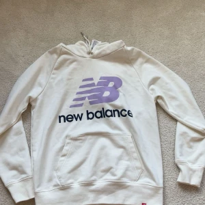 Vit hoodie från New Balance S - Snygg vit hoodie från New Balance i storlek S med lila logga framtill och svart text. Tröjan har huva med snörning och en stor magficka. Perfekt för en chill och sportig look. Tillverkad i mjuk bomullsmix som känns skön mot huden.
