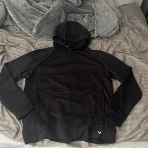 Svart hoodie från SOC i polyester - Svart hoodie från SOC i storlek M, tillverkad i teknisk polyester med TCS Airflow för bra ventilation. Tröjan har huva, långa ärmar och en diskret logga framtill. Perfekt för chill dagar.