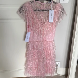 Ljusrosa spetsklänning med volanger - Supersöt ljusrosa kortklänning från BubbleRoom i spets med flera lager volanger och romantiska detaljer. Klänningen har ärmlös design, v-ringning och ett feminint, fluffigt uttryck. Perfekt för dig som vill sticka ut med en drömmig look.