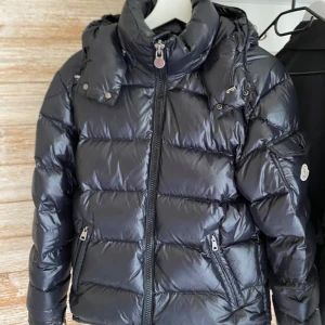 Moncler Maya Giubbotto  - Säljer en svart Moncler Maya Giubbotto. Jackan har dragkedja framtill, logga på ärmen och coolt innerfoder med serietidningsmönster. Perfekt för kalla dagar och riktigt snygg.  Vid frågor eller flera bilder så är det bara att skriva!  Fick inte plats med alla bilder här. Jackan har storlek 1 alltså S och är killmodell men jag som tjej har använt den och ser inga problem med den utan att den var för stor för mig.