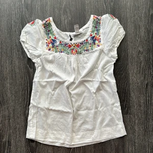 Vit broderad blus från H&M - Superfin vit blus från H&M med färgglad brodyr upptill och på axlarna. Blusen har korta puffärmar och rund halsringning. Broderiet har blommor och fågelmotiv i rött, blått, grönt, gult och lila. Luftig och skön i bomull, perfekt för vår och sommar.