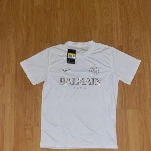Vit PSG x Balmain fotbollströja - Snygg vit fotbollströja från Nike i samarbete med PSG och Balmain. Tröjan har korta ärmar, rund hals och holografiskt tryck med Balmain Paris-logga samt PSG-emblem på bröstet. Perfekt för dig som gillar exklusiva samarbeten och stilren design.