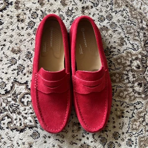 Röda mockaloafers från Rockport - Snygga röda loafers från Rockport i mjuk mocka med klassisk pennyloafers-design. Skorna har en platt sula och rund tå, samt bekväm innersula. Perfekta för dig som vill sticka ut med färg och stil. Ordinarie pris: 1300kr