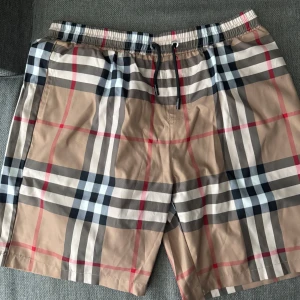 Burberry rutiga shorts med snörning - Snygga shorts från Burberry i klassiskt beige, svart, vit och röd rutig design. De har elastisk midja med svart snörning och är tillverkade i ett lätt syntetmaterial. Perfekta för sommaren och ger en clean och trendig vibe. Storlek passar XL och L