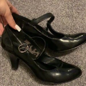 Svarta pumps med rem och klack - Snygga svarta pumps från Chilli med rund tå, klassisk T-rem och spänne. Skorna har en medelhög klack och är tillverkade i glansigt syntetmaterial. Perfekta för dig som vill ha en stilren och feminin look med extra detalj på foten.