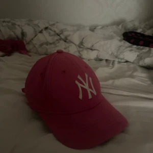 Rosa New York Yankees keps - Snygg rosa keps från New York Yankees med vit broderad NY-logga framtill. Klassisk böjd skärm och justerbar passform. Perfekt accessoar för att lyfta din outfit med lite färg och sportig vibe.